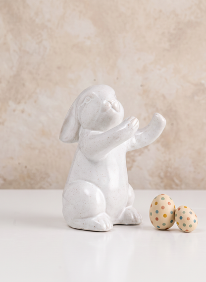 A 6.5in Aged White Porcelain Rabbit from a set of four is displayed sitting upright beside two pastel, polka-dotted eggs on a white surface, with a light beige textured background.