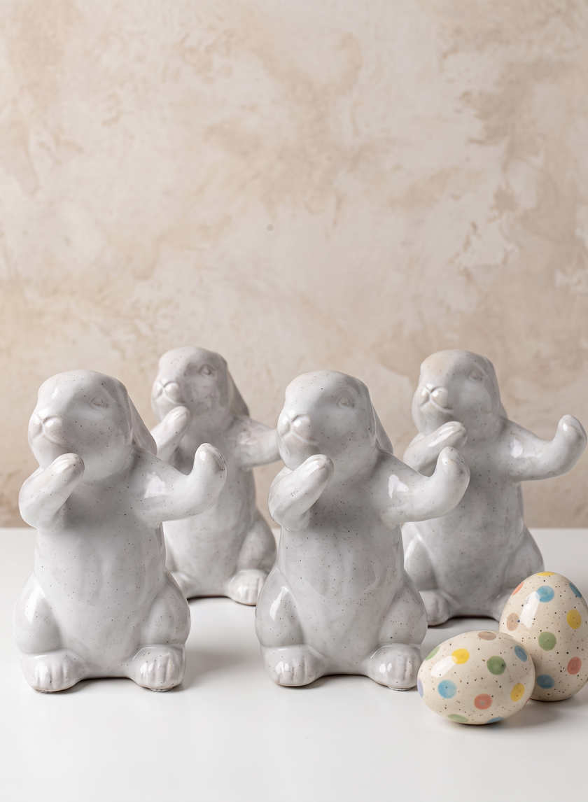 A set of four 6.5in Aged White Porcelain Rabbits stand together beside two pastel speckled Easter eggs on a white surface, against a beige textured background.
