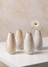 Four "Travertine Look" 5-inch tall ceramic bud vases in beige are arranged on a white table; one vase displays a single white daisy with a yellow center, while the remaining three are empty, set against a neutral background.