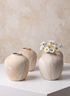 Four round, beige "Travertine Look" ceramic bud vases (4in tall) are arranged on a white surface. Three of the vases are empty, while one features a bouquet of small white daisies. The background is a light, textured wall.