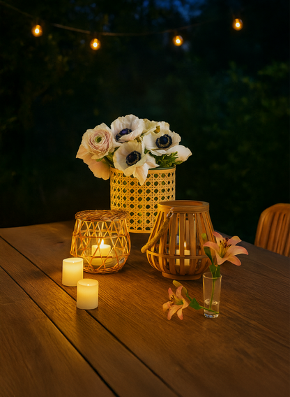 A wooden outdoor table with lit candles, a vase of white flowers, a small glass with peach lilies, and two Indochine Cane Weaved Glass Cylinders as lanterns. String lights glow in the night sky background.