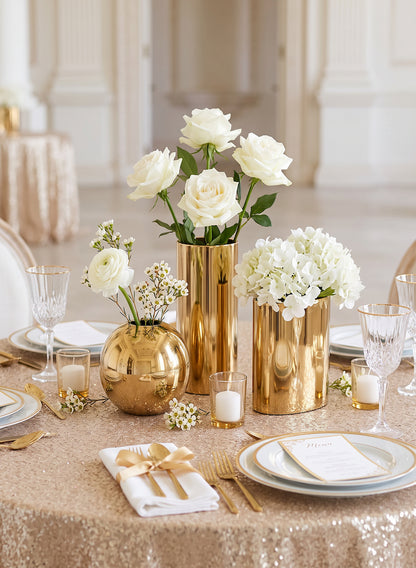 Elegant table setting with gold vases, white roses, and hydrangeas on a sparkling tablecloth, surrounded by glassware and
