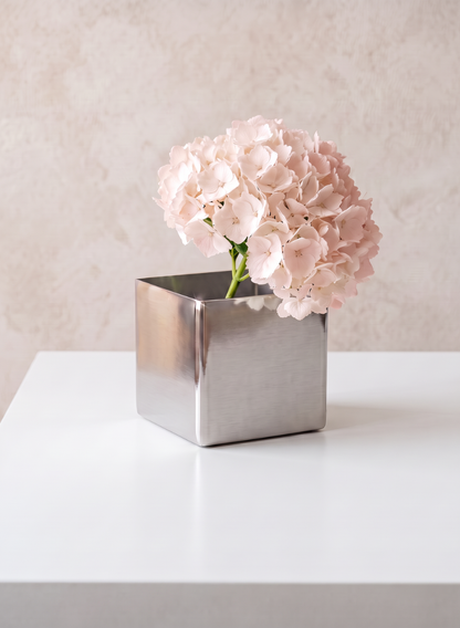 A light pink hydrangea flower in a modern, 3in Cube Polish Stainless Steel Vase sits on a white table against a light beige textured background.