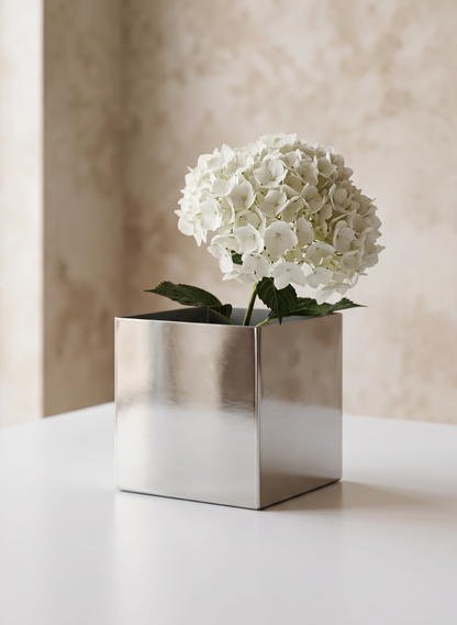 A white hydrangea with green leaves is arranged in a 5in Cube Polish Stainless Steel Vase, set on a white surface with a beige textured wall in the background.