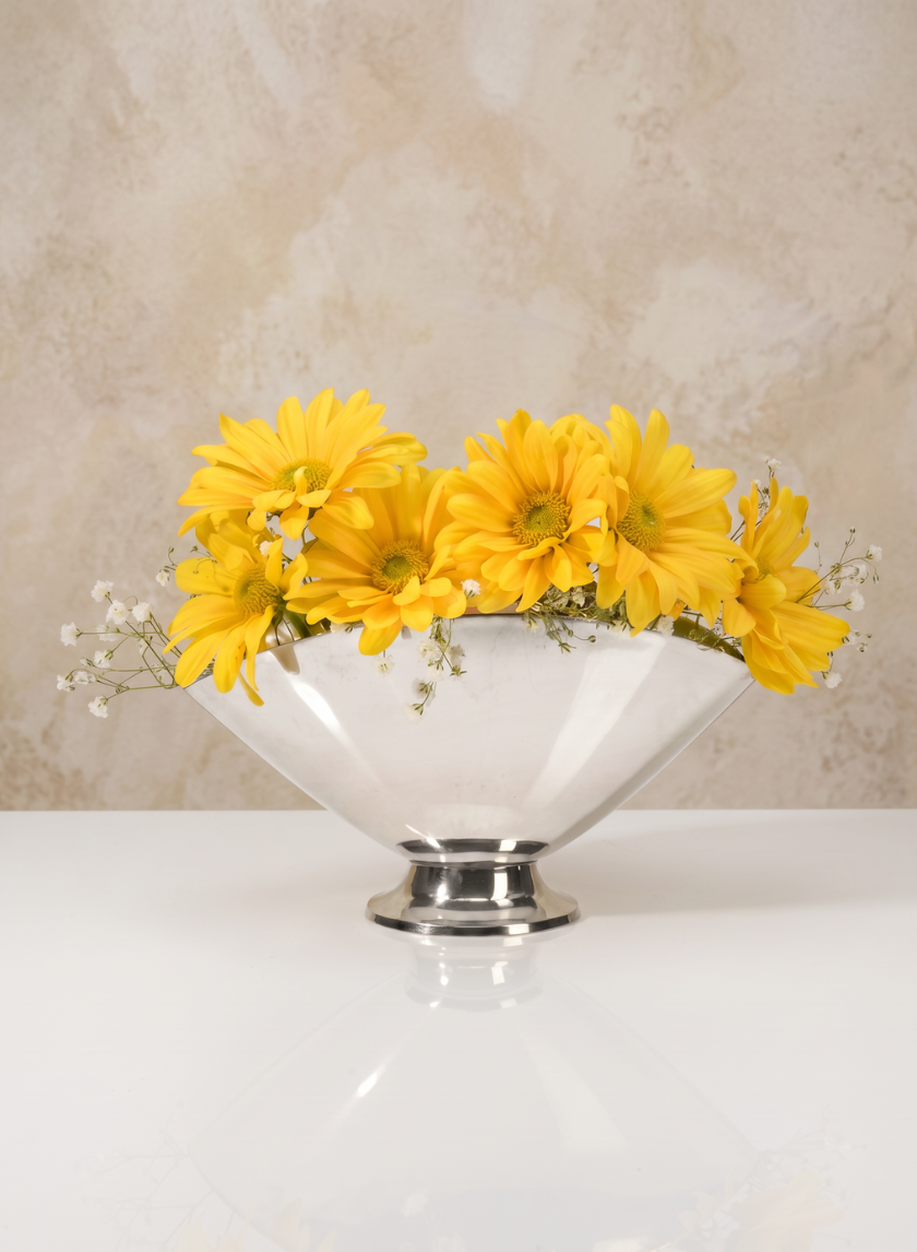 A 6.5in Wide Nickel Oval Bowl holds bright yellow daisies and small white flowers, set on a reflective white surface with a beige textured background.