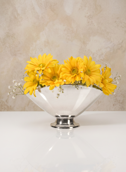 A 6.5in Wide Nickel Oval Bowl holds bright yellow daisies and small white flowers, set on a reflective white surface with a beige textured background.