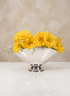 A 6.5in Wide Nickel Oval Bowl holds bright yellow daisies and small white flowers, set on a reflective white surface with a beige textured background.