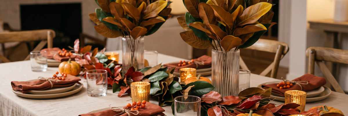 An autumn-themed dining table decorated with a green and red leaf garland, large brown magnolia leaf arrangements in glass vases, lit gold pillar candles, and plates with folded brown napkins.