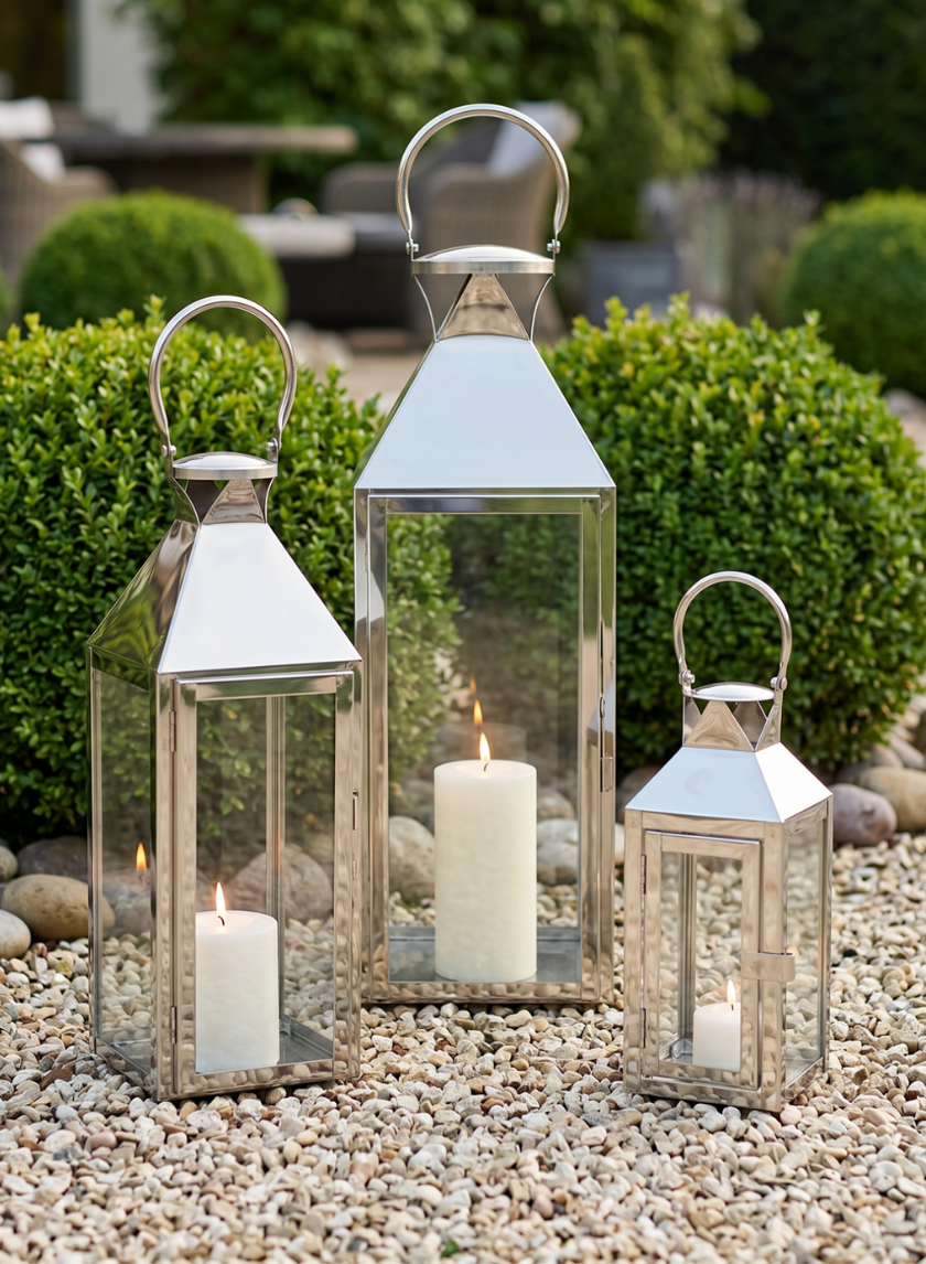 Three Silver Soho Stainless Steel Lanterns with white pillar candles inside sit on light-colored pebbles, green-trimmed bushes and outdoor furniture blurred behind—an elegant choice for wedding lanterns.