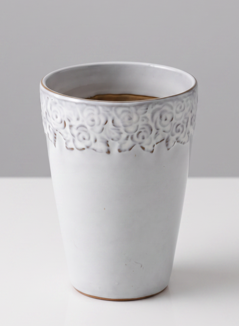 A 6in tall Aurelie Rose Border Cachepot, featuring a white ceramic finish with an embossed floral design near the rim, is shown filled with a brown liquid and placed on a white surface against a light gray background.