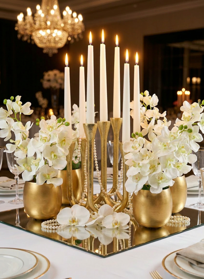 Elegant table centerpiece featuring tall white candles in a gold holder, surrounded by lush 33in Long Green Double Orchid Stems (case of 12) arranged in gold vases on a mirrored base. Chandeliers and glassware complete the luxurious wedding décor and formal setting.